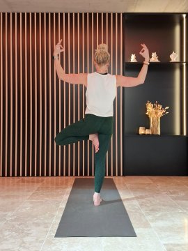 Frau macht Yoga im Baum-Pose auf Matte vor Wand mit vertikalen Holzleisten und Regal