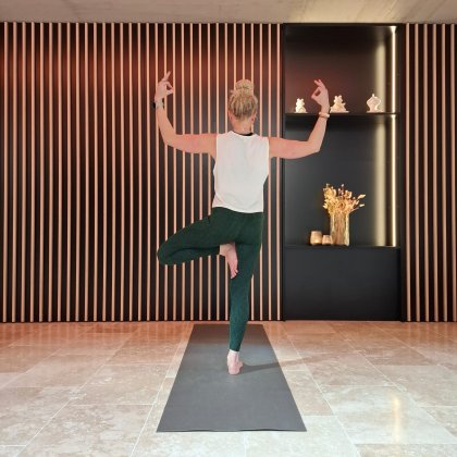 Frau macht Yoga im Baum-Pose auf Matte vor Wand mit vertikalen Holzleisten und Regal