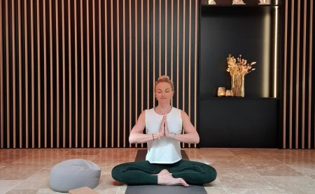 Frau sitzt im Schneidersitz auf Yogamatte, Hände in Gebetshaltung, vor Wand mit Holzleisten