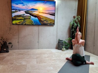 Person macht Yoga auf Matte rechts im Raum mit Pflanzen und Fernseher an der Wand