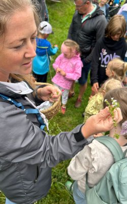 Frau zeigt Kindern im Freien eine kleine Blume, umgeben von mehreren Kindern und Erwachsenen