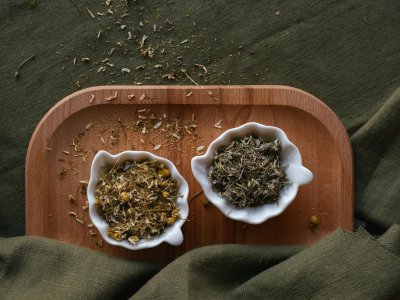 Zwei weiße Schalen mit getrockneten Kräutern auf einem Holzbrett, umgeben von grünem Stoff
