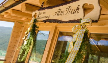 Holzschild „AlmRuh“ mit Herzen, Schleifen und Tannenzweigen