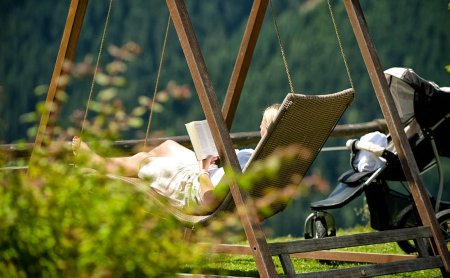 Frau liest auf einer Gartenschaukel mit Kinderwagen im Hintergrund