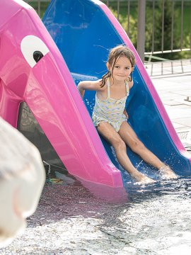 Mädchen rutscht lachend auf pink-blauer Rutsche ins Kinderbecken