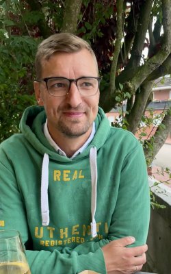 Mann mit Brille und grünem Hoodie sitzt draußen an einem Tisch vor Bäumen