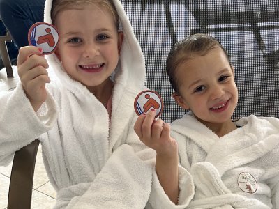 Zwei lächelnde Kinder in weißen Bademänteln sitzen nebeneinander und halten Abzeichen hoch