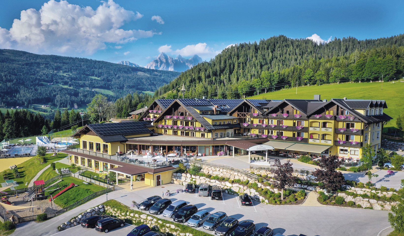 Hotel mit gelber Fassade, Blumendekor und Bergpanorama im Hintergrund