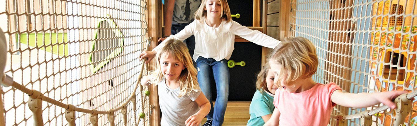 Kinder balancieren lachend über eine Hängebrücke im Indoor-Spielbereich