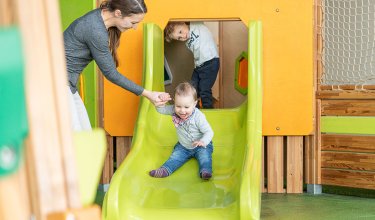 Kleinkind rutscht mit Unterstützung der Mutter die grüne Rutsche hinunter