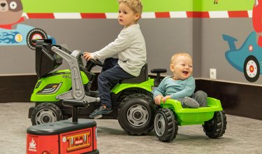 Zwei Kleinkinder spielen mit grünem Trettraktor und Anhänger