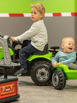 Zwei Kleinkinder spielen mit grünem Trettraktor und Anhänger