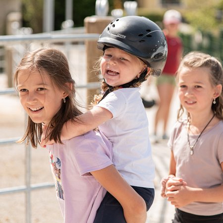 Ein Kind trägt ein anderes Kind auf dem Rücken, beide vor einem Reitstall.