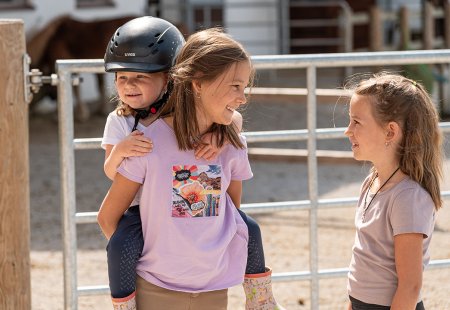 A child on another child’s back in front of a riding stable, both wearing T-shirts.