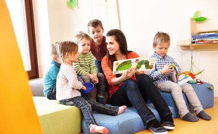 Erzieherin liest mit Kindern ein Bilderbuch in einem hellen Spielraum.
