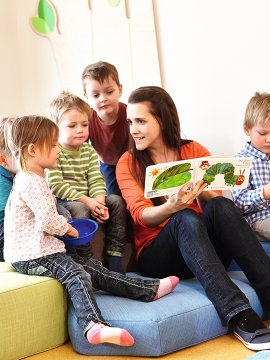 Erzieherin liest mit Kindern ein Bilderbuch in einem hellen Spielraum.