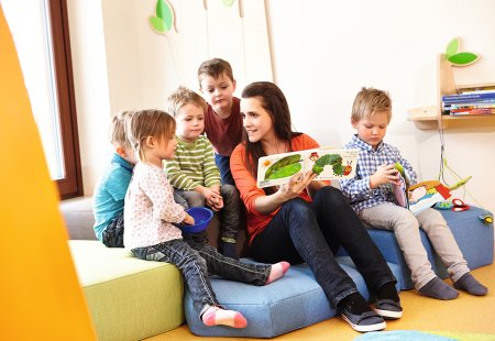 Erzieherin liest mit Kindern ein Bilderbuch in einem hellen Spielraum.