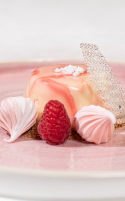 Dessert mit Himbeeren, Baiser und Zuckerhülle auf einem rosa Teller.