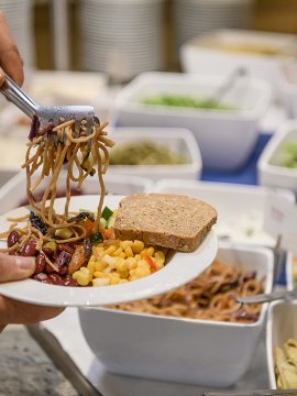 Hand nimmt mit Gabel Nudeln vom Buffet auf einen Teller mit Brot, Mais und Bohnen