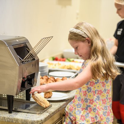 Mädchen steckt Brot in Toaster in der Küche, im Hintergrund steht ein weiteres Kind mit Teller