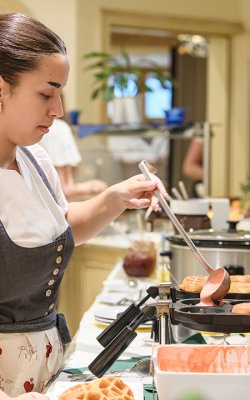 Frau gießt Teig in Herzwaffeleisen in einer Küche mit Pflanzen und Spiegel im Hintergrund