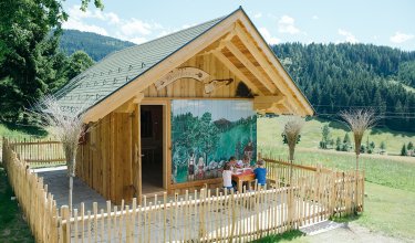 Bunt bemaltes Holzhaus mit umzäuntem Außenbereich, Kinder malen an einem Tisch.