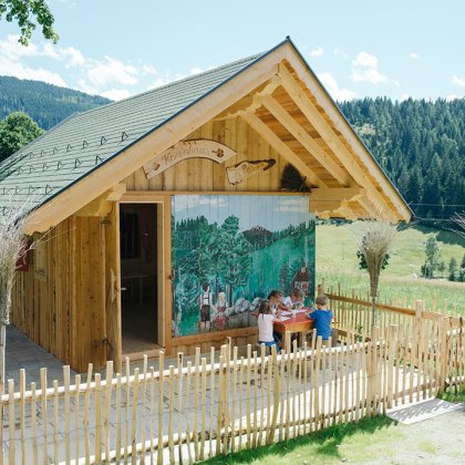 Bunt bemaltes Holzhaus mit umzäuntem Außenbereich, Kinder malen an einem Tisch.
