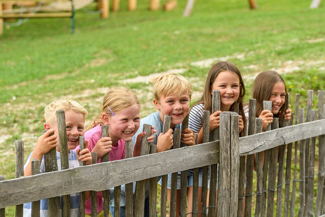 Fünf lachende Kinder lehnen sich hinter einem Holzzaun im Freien
