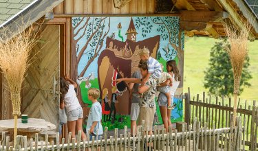 Kinder schauen und zeigen auf ein buntes Wandbild an einem Holzhaus mit Zaun und ländlicher Umgebung