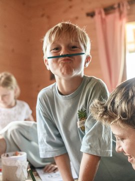 Children sitting at a table in a wooden room, one boy making a face with a rubber band