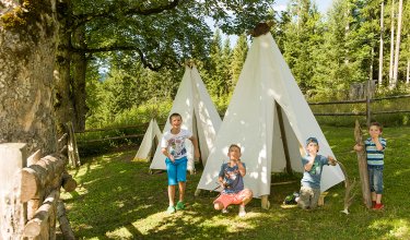 Vier Kinder spielen vor weißen Tipis im Freien, umgeben von Bäumen.