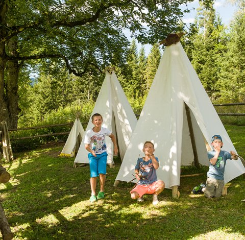Vier Kinder spielen vor weißen Tipis im Freien, umgeben von Bäumen.