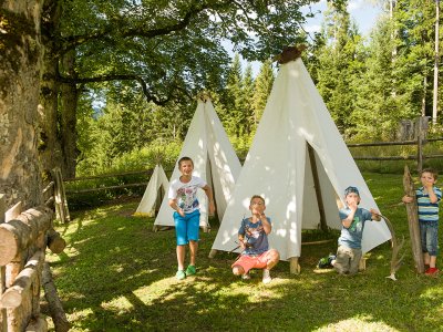 Vier Kinder spielen vor weißen Tipis im Freien, umgeben von Bäumen.