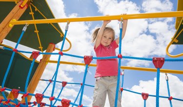 Ein Mädchen klettert an einem Seilnetz auf einem Spielplatz, mit blauem Himmel und Wolken im Hintergrund.