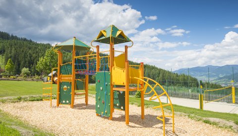 Ein bunter Spielplatz mit Klettergerüst und Rutsche, umgeben von grüner Wiese und Bergen.