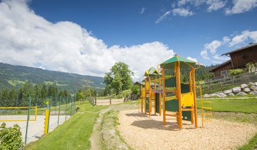 Ein Spielplatz mit Klettergerüst und Rutsche, umgeben von grüner Wiese und Bergen.