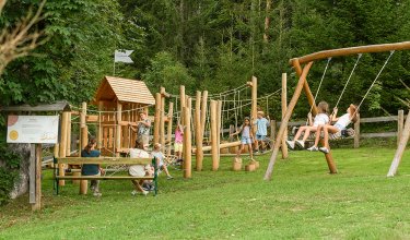 Children playing on a wooden playground with swings and climbing structures, adults sitting on benches