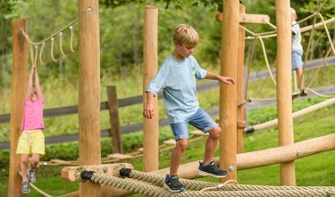 Kind balanciert auf einem dicken Seil im Kletterpark mit weiteren Kindern im Hintergrund