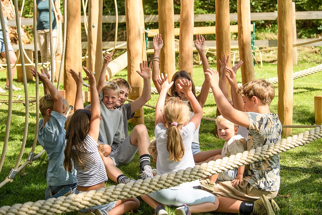 Kinder sitzen im Kreis auf Gras im Freien und heben die Arme spielerisch hoch
