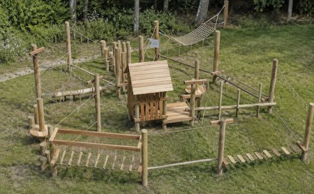 Holz-Klettergerüst mit Hängebrücken und Netz in einem grasbewachsenen Bereich neben Bäumen