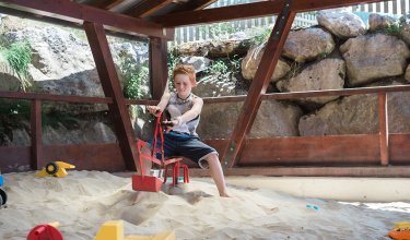 Junge sitzt im Sandkasten und spielt mit einem roten Schaufelbagger.