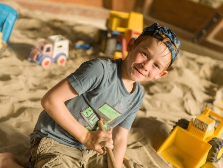 Junge im Sandkasten spielt mit einem Schaufelbagger und lächelt in die Kamera.