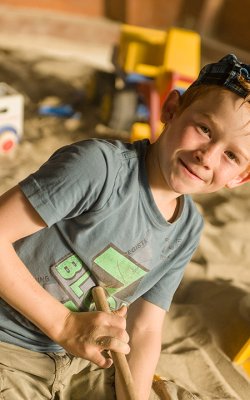 Junge im Sandkasten spielt mit einem Schaufelbagger und lächelt in die Kamera.