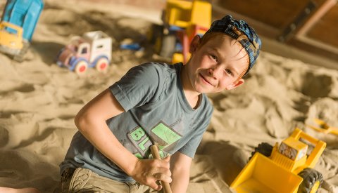 Junge im Sandkasten spielt mit einem Schaufelbagger und lächelt in die Kamera.