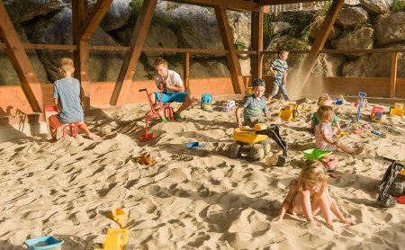 Kinder spielen im Sandkasten mit Baggern und Autos unter einem Holzüberdachung.