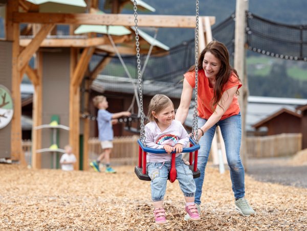 Frau schiebt lachendes Kind auf Schaukel auf einem Spielplatz mit Holzhäusern im Hintergrund
