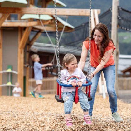 Frau schiebt lachendes Kind auf Schaukel auf einem Spielplatz mit Holzhäusern im Hintergrund