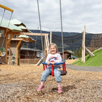 Two children swinging on a playground with wooden structures and mountains in the background