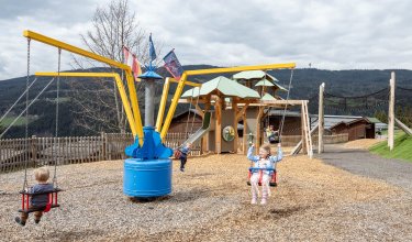Kinder schaukeln auf einem Spielplatz mit gelb-blauem Karussell und Holzhütte im Hintergrund
