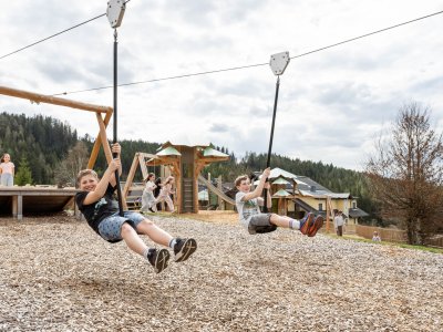 Zwei Jungen fahren auf einer Seilrutsche auf einem Spielplatz, im Hintergrund spielen Kinder und Bäume